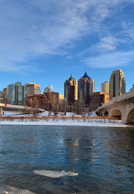 A cityscape view featuring a river in the foreground with dark, slightly rippled water and patches of ice along the edges. On the riverbank, there is a narrow strip of snow-covered ground with bare de