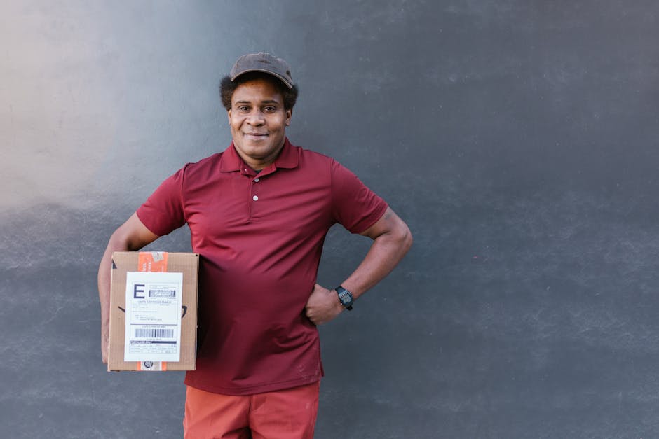 A delivery person with medium brown skin and short curly hair, wearing a gray cap, maroon polo shirt, and red pants, stands in front of a dark gray wall. He is holding a medium-sized, sealed cardboard